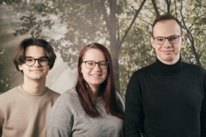 Gruppenfoto. 2 junge, weiße Männer mit Brille und in der Mitte eine junge weiße Frau mit Brille.